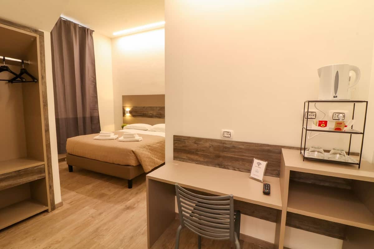 A hotel room with a bed, a desk, a chair, a wardrobe, and a coffee machine.