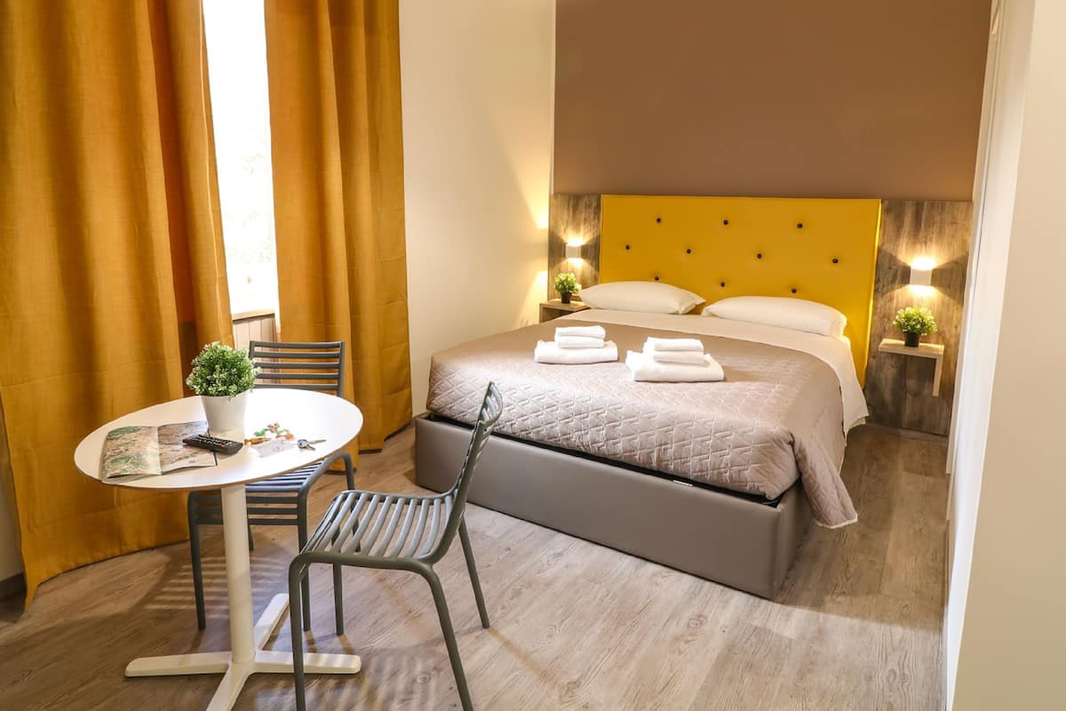 A hotel room with a bed, a small table with a plant, a chair, and a bedside table with a lamp.