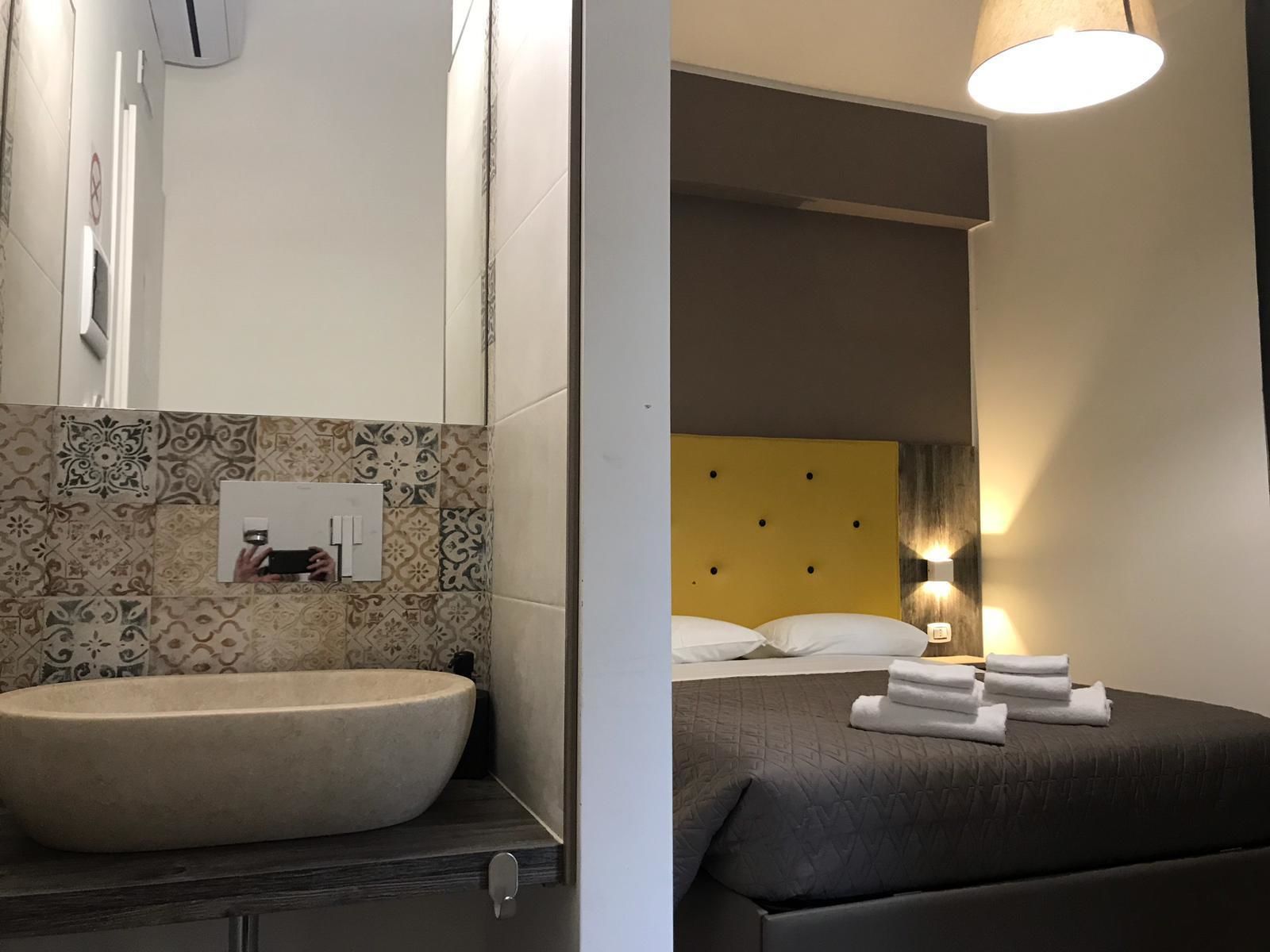 Un bagno con un lavandino grande e uno specchio, e una camera da letto con un letto, cuscini e una lampada da comodino.