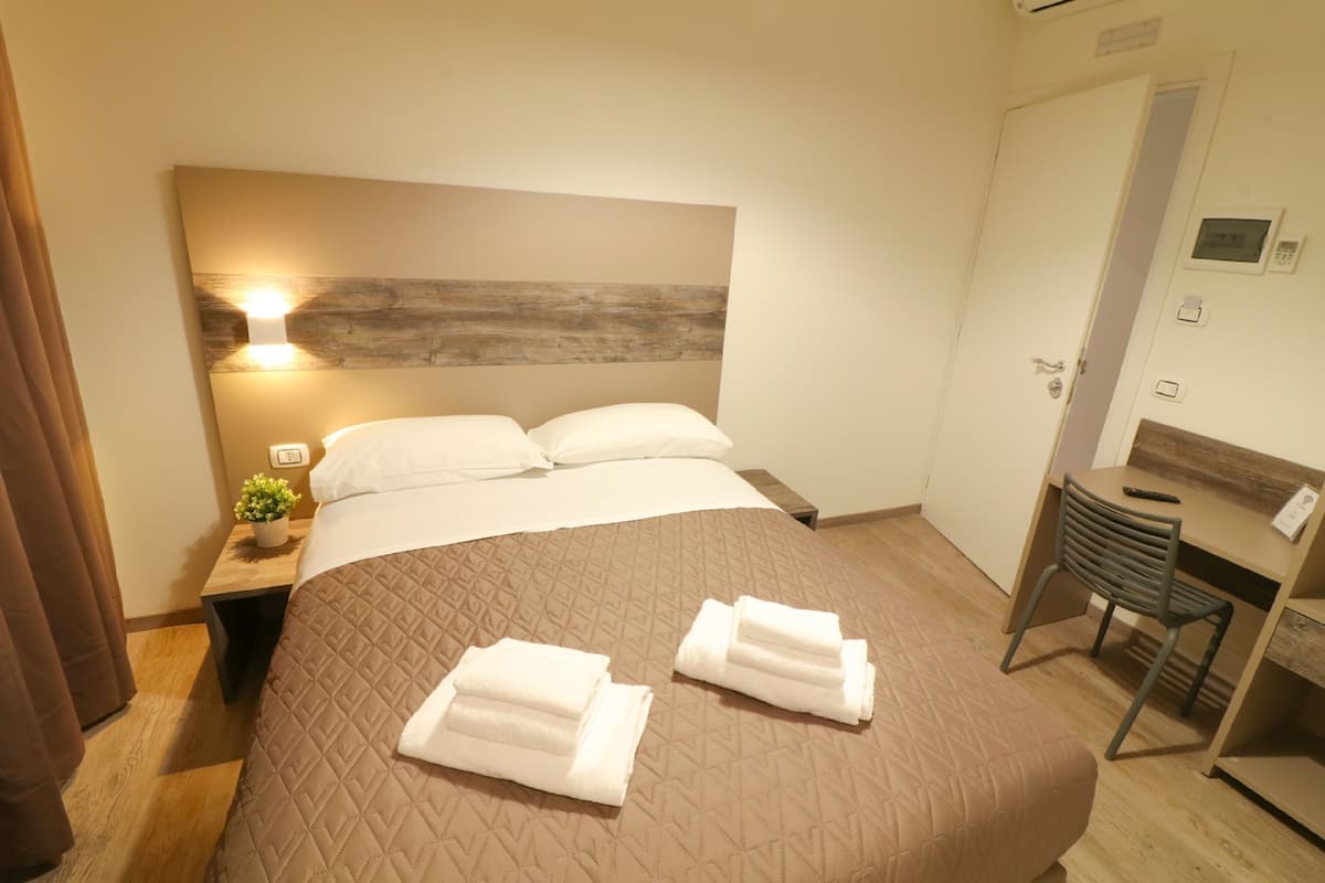 A neatly made bed with two folded towels, a bedside table with a plant, and a desk with a chair.