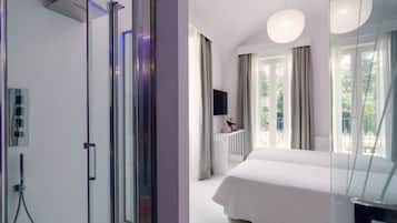 Moderni hotellihuone, jossa on suuri sänky, lasinen suihkuseinä, televisio ja parveke, jossa on verhot.