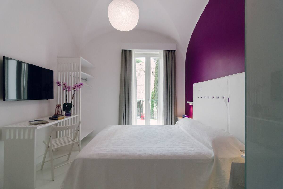 A bedroom with a bed, a desk, a chair, a TV, and a window with curtains.