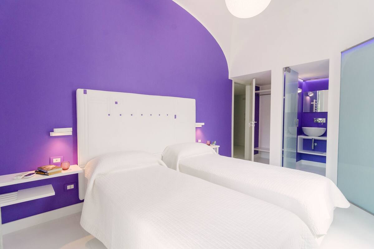 A modern bedroom with two beds, a purple accent wall, a white nightstand, and a bathroom with a blue accent wall.