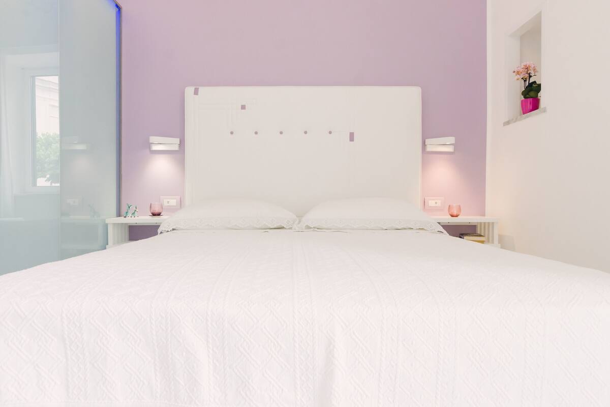 A neatly made bed with a white headboard, two bedside tables with lamps, and a small window with a potted plant.
