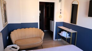 Une chambre d’hôtel avec un canapé beige, une petite table de chevet avec une télécommande, un miroir et une étagère avec des serviettes.