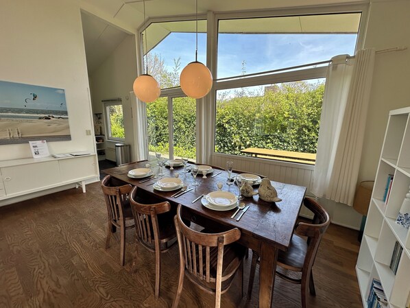 A dining area with a large table set for four, surrounded by wooden chairs, large windows with a view of greenery, and pendant lighting.