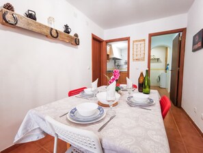 Un coin repas avec une table dressée pour quatre personnes, comprenant des assiettes, des verres et des couverts. On y trouve une chaise rouge et une chaise blanche avec un dossier. Une étagère en bois, située au-dessus de la table, expose des objets décoratifs.