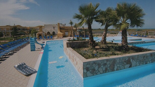 A swimming pool area with blue water, lounge chairs, palm trees, and a stone wall. - Villaggio Turistico Le Meridiane (Montenero di Bisaccia)