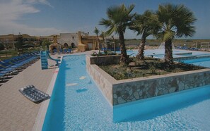 Un espace piscine avec de l’eau bleue, des chaises longues, des palmiers et un mur en pierre.