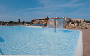 Una piscina con fuente de agua, sillas de descanso y edificios al fondo.