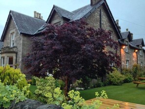 A stone building with a sloped roof and a well-maintained garden with a variety of plants and a bench. - Buttonboss Lodge (Pitlochry)