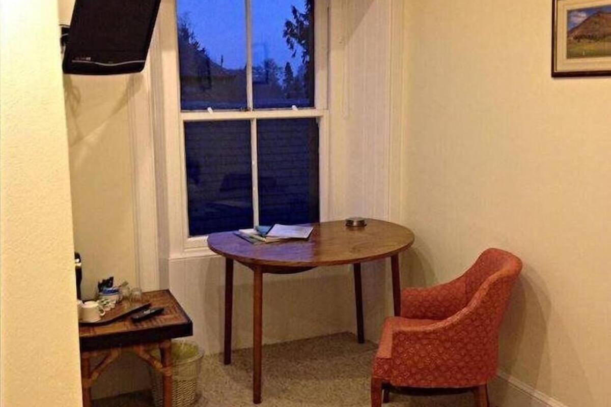 A small, cozy room with a single round table, a red chair, a television mounted on the wall, and a window with a view of trees.