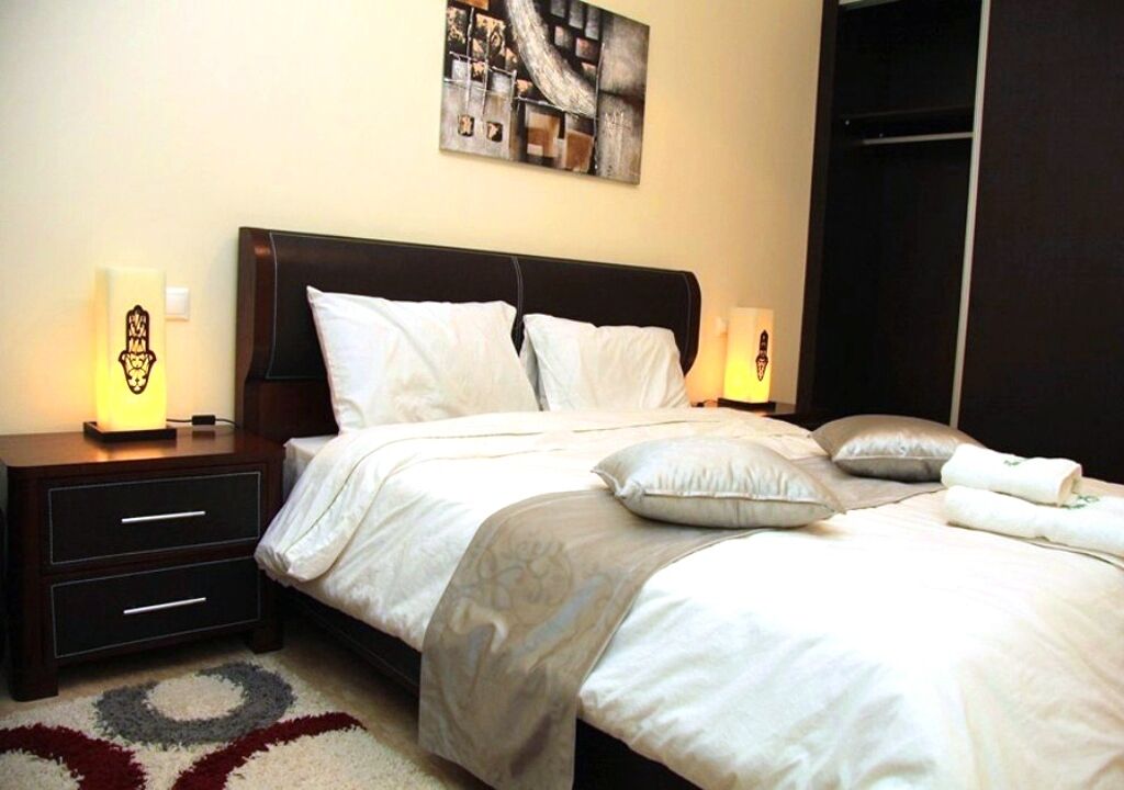Una cama bien hecha con ropa de cama blanca y un plaid gris, una mesita de noche de madera oscura con una lámpara, y un cuadro abstracto moderno en la pared.
