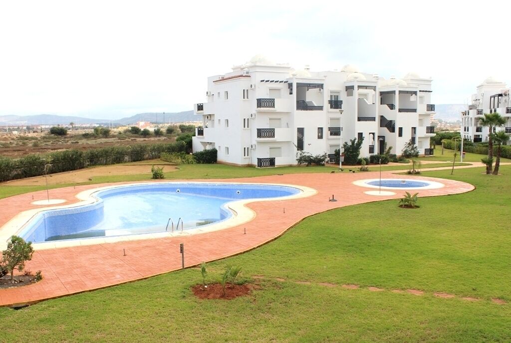 Un moderno edificio residenziale con numerosi balconi, un giardino curato con piscina e vista sulle montagne in lontananza.