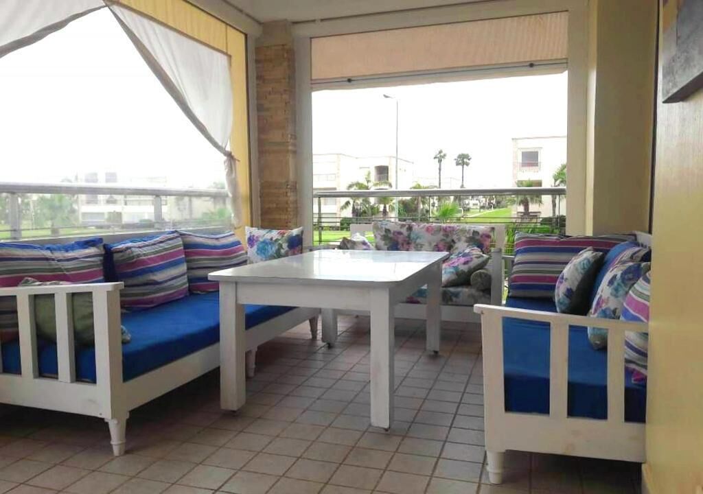 Ein Zimmer mit einem weißen Sofa und einem weißen Tisch, beide mit blauen Kissen. Ein großes Fenster bietet einen Blick nach draußen.