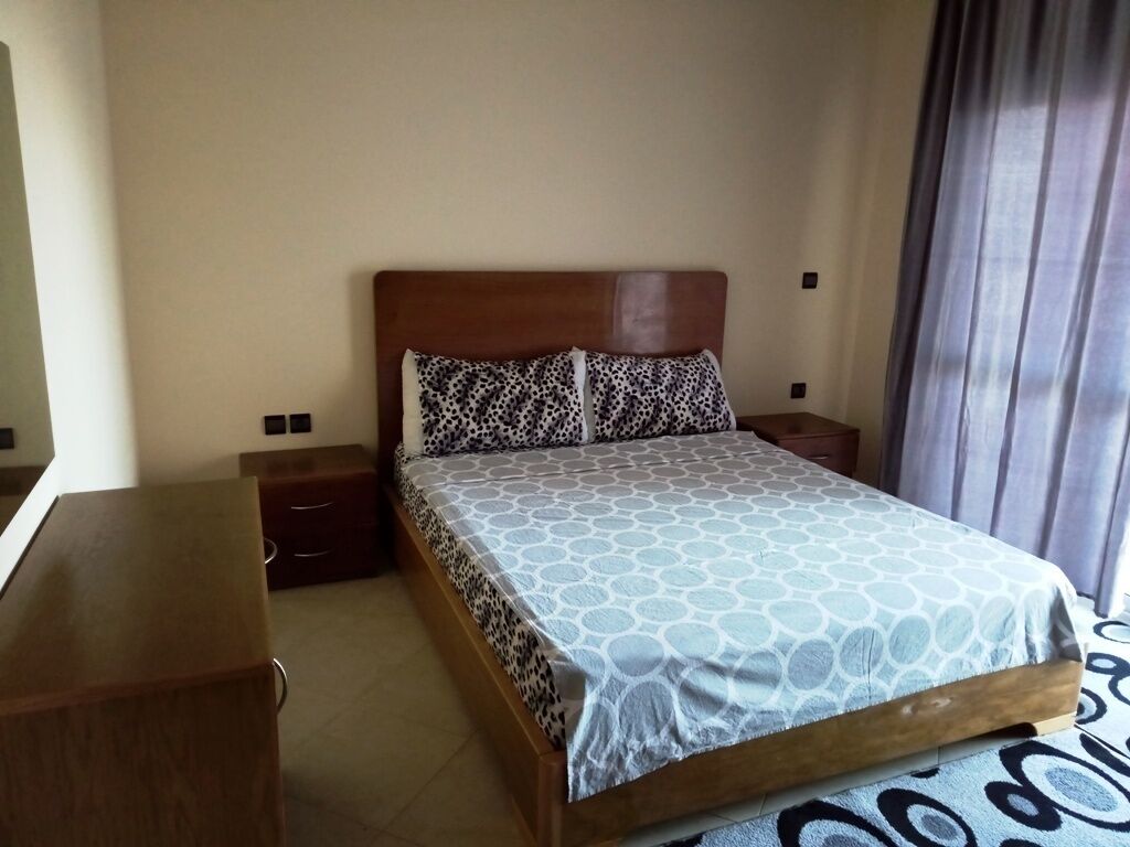 A hotel room with a bed, a wooden nightstand, a dresser, and a window with curtains.
