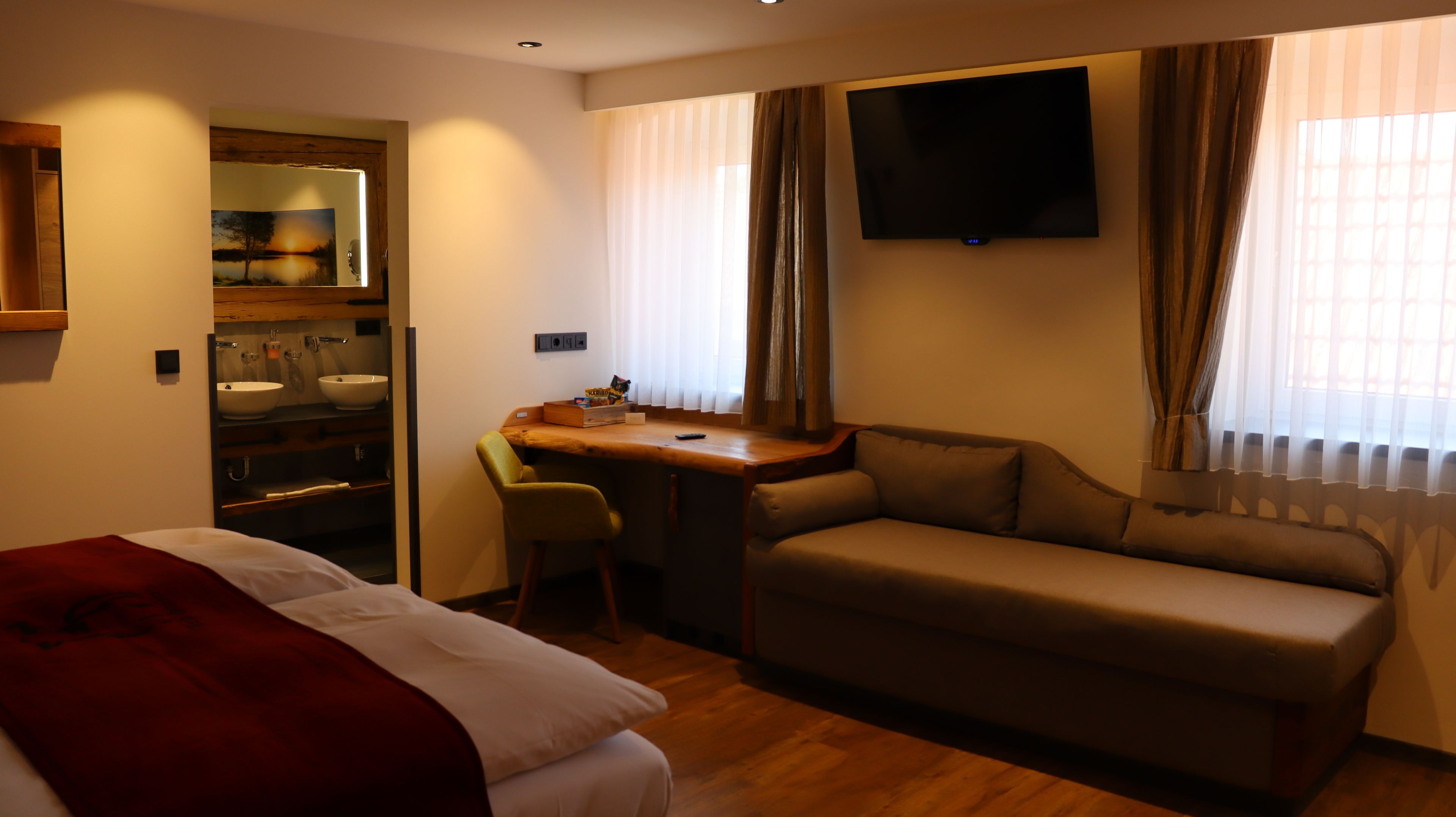 A hotel room with a bed, a desk, a sofa, a TV, and a bathroom with a sink and mirror.