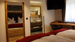Une chambre avec un lit en bois, un bureau, une télévision et une salle de bain équipée d’un miroir et d’un lavabo.