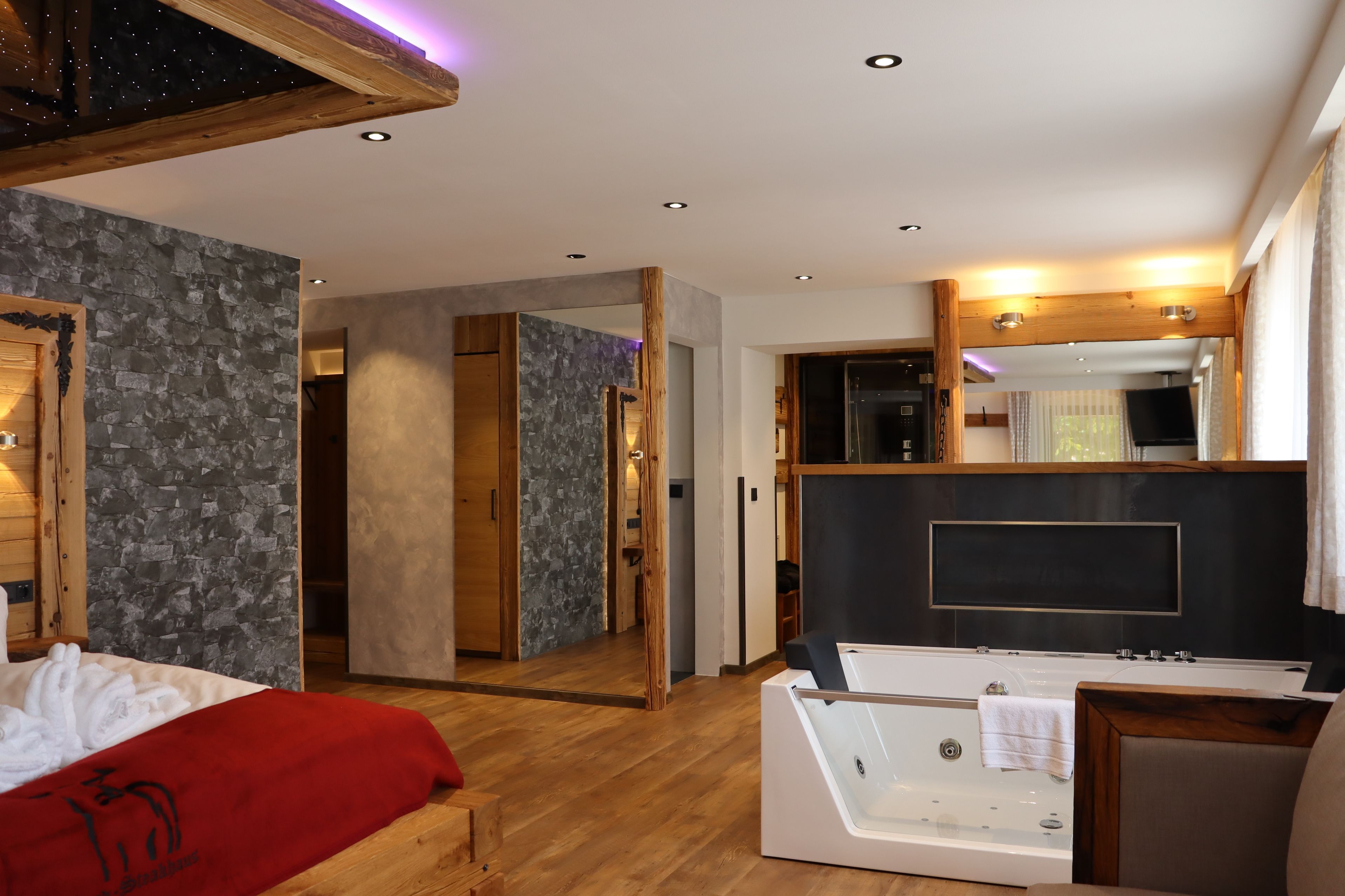 Habitación de hotel moderna con suelo de madera, pared de acento de piedra, espejo grande, bañera y colcha roja.