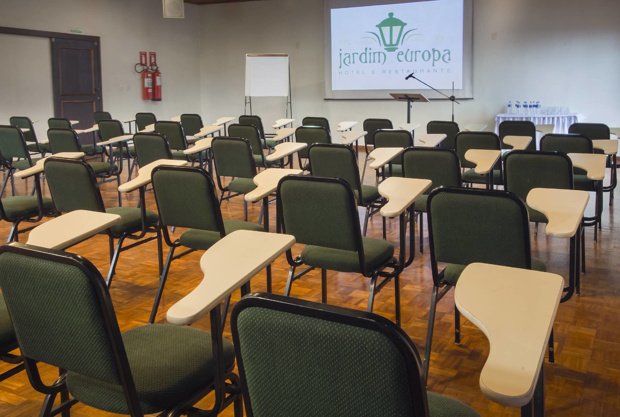 Uma sala de conferências com cadeiras verdes e mesas brancas, uma lousa, um projetor com o logotipo 'jardin europa hotel e restaurante' e extintores na parede.