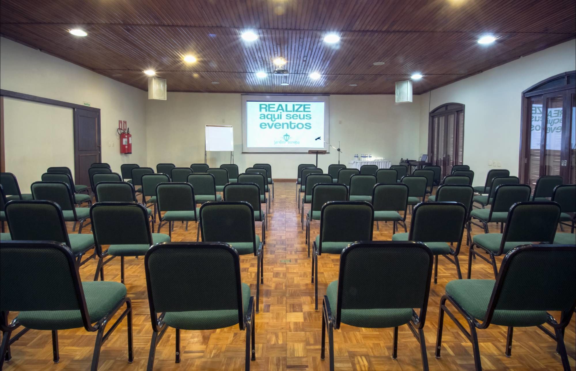 Un salón de conferencias con filas de sillas verdes, piso de madera y una pantalla proyectando texto.