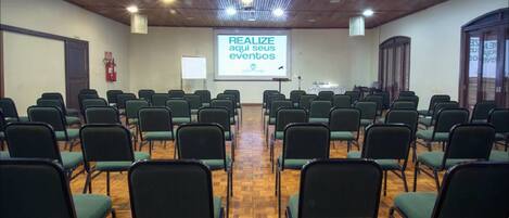Un salón de conferencias con filas de sillas verdes, piso de madera y una pantalla proyectando texto.