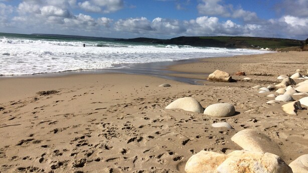 Una playa arenosa con rocas grandes e huellas, cielo despejado y olas rompiendo en la orilla.