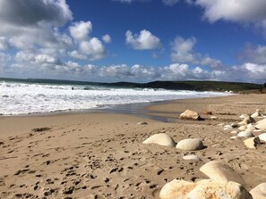 Una playa arenosa con rocas grandes e huellas, cielo despejado y olas rompiendo en la orilla.