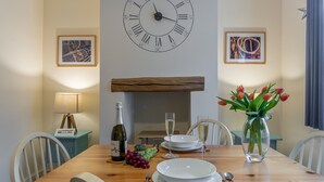 Ein Esstisch ist gedeckt mit Tellern, Besteck, einer Flasche und einem Blumenstrauß. An der Wand über dem Tisch hängt eine Uhr.