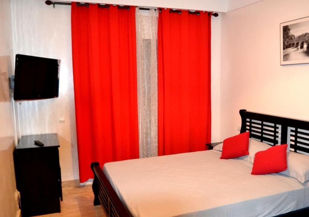A bedroom with a bed, red curtains, a small table, and a mounted TV.