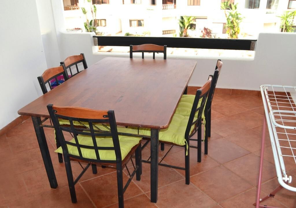 A dining area with a wooden table and chairs, a metal rack, and a view of buildings and plants through a window.
