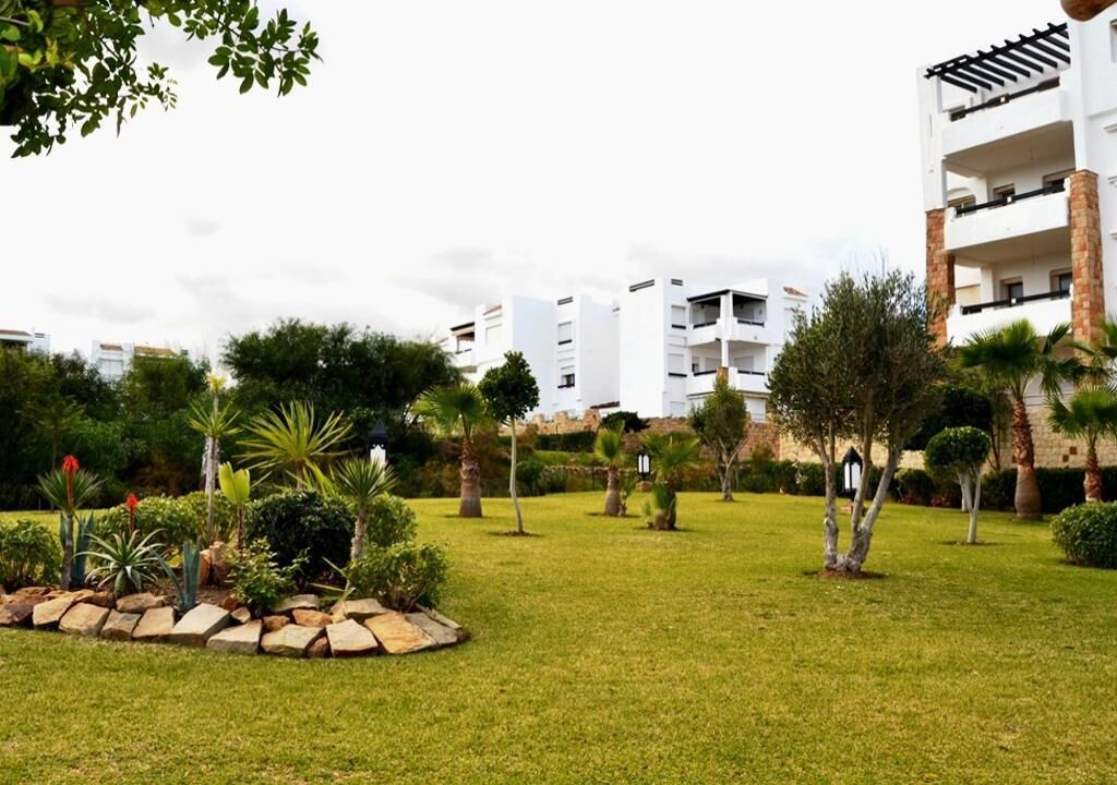 A residential area with modern white buildings, a well-maintained grassy lawn, and various trees and shrubs.