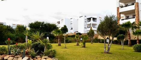 A residential area with modern white buildings, a well-maintained grassy lawn, and various trees and shrubs.