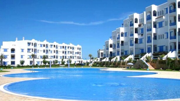 Ein Resort mit großem Schwimmbecken, weißen Gebäuden mit blauen Akzenten und Palmen.
