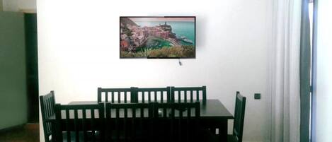 A dining area with a table and chairs, an air conditioning unit, and a painting of a coastal town on the wall.