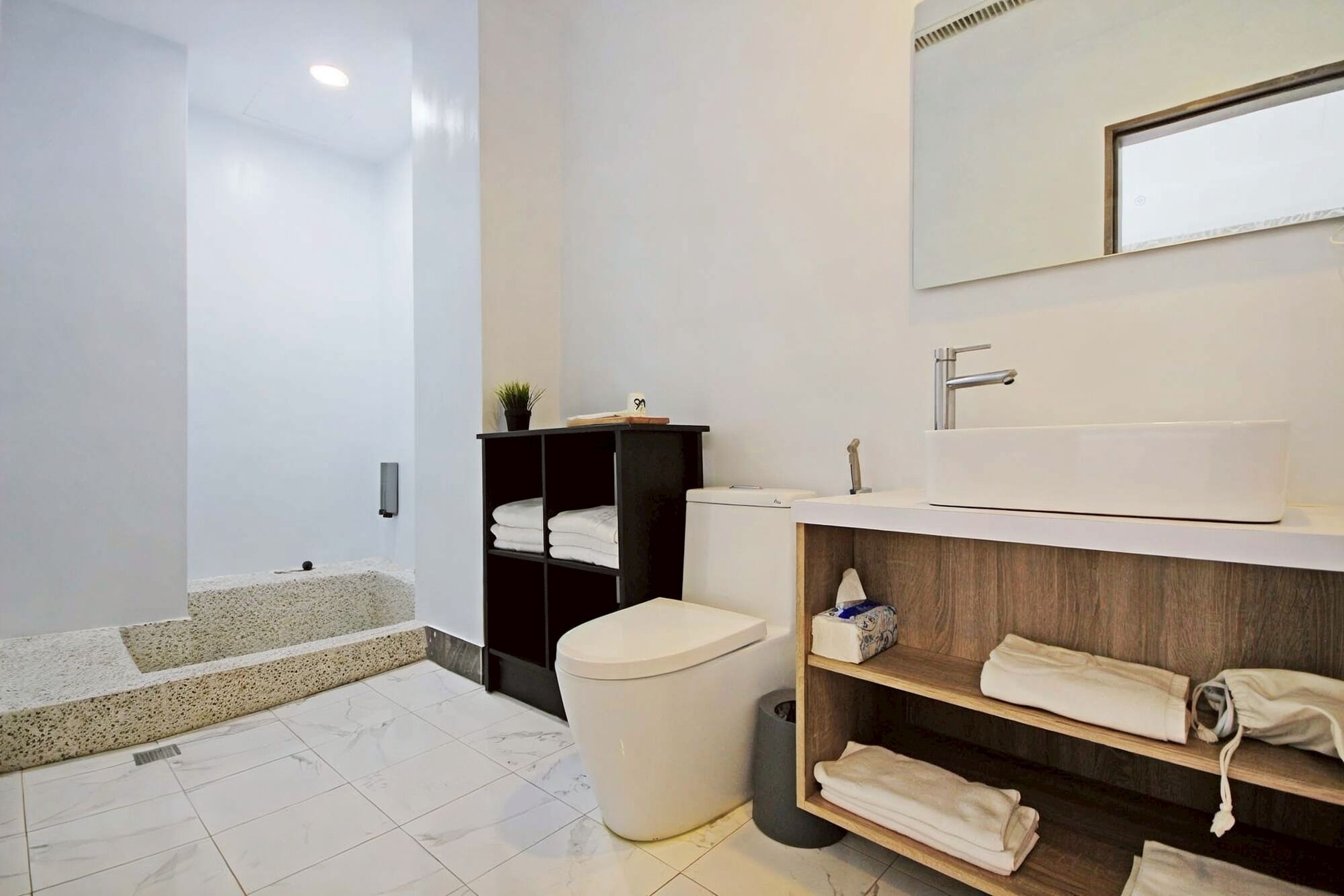 Un baño moderno con inodoro blanco, lavamanos con estante de madera y zona de ducha.