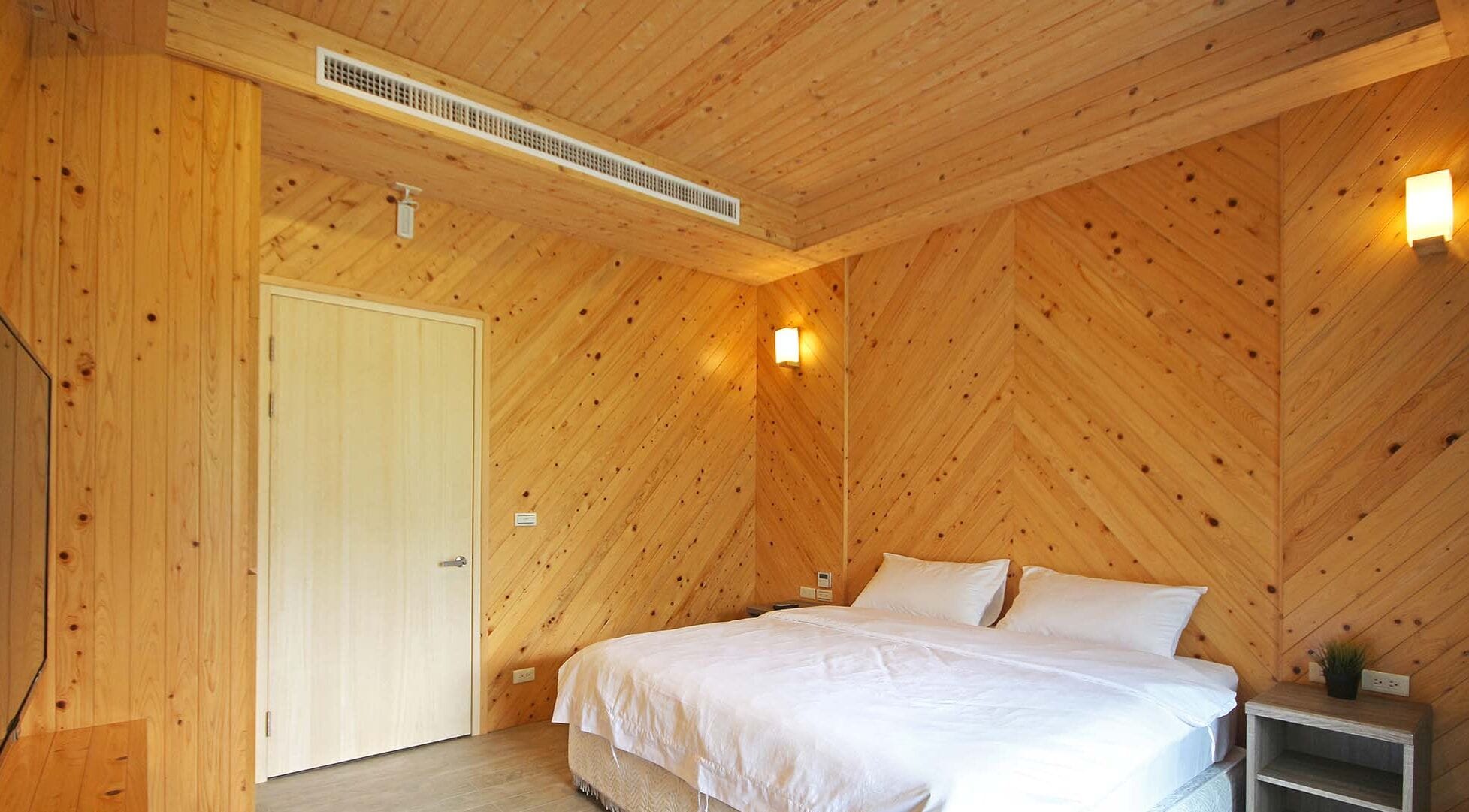 A bedroom with a wooden sloped ceiling, a bed with white bedding, a bedside table, and a wall-mounted light fixture.