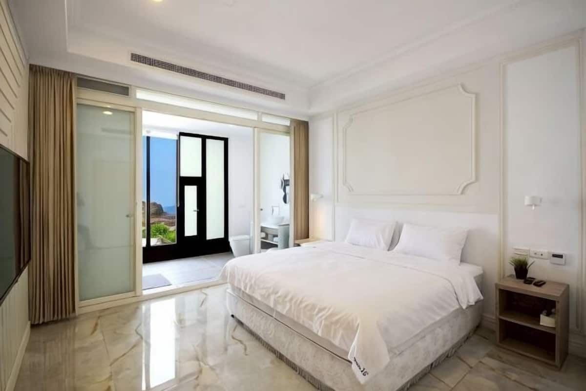 A hotel room with a large bed, a desk, a chair, and a sliding glass door leading to a balcony.
