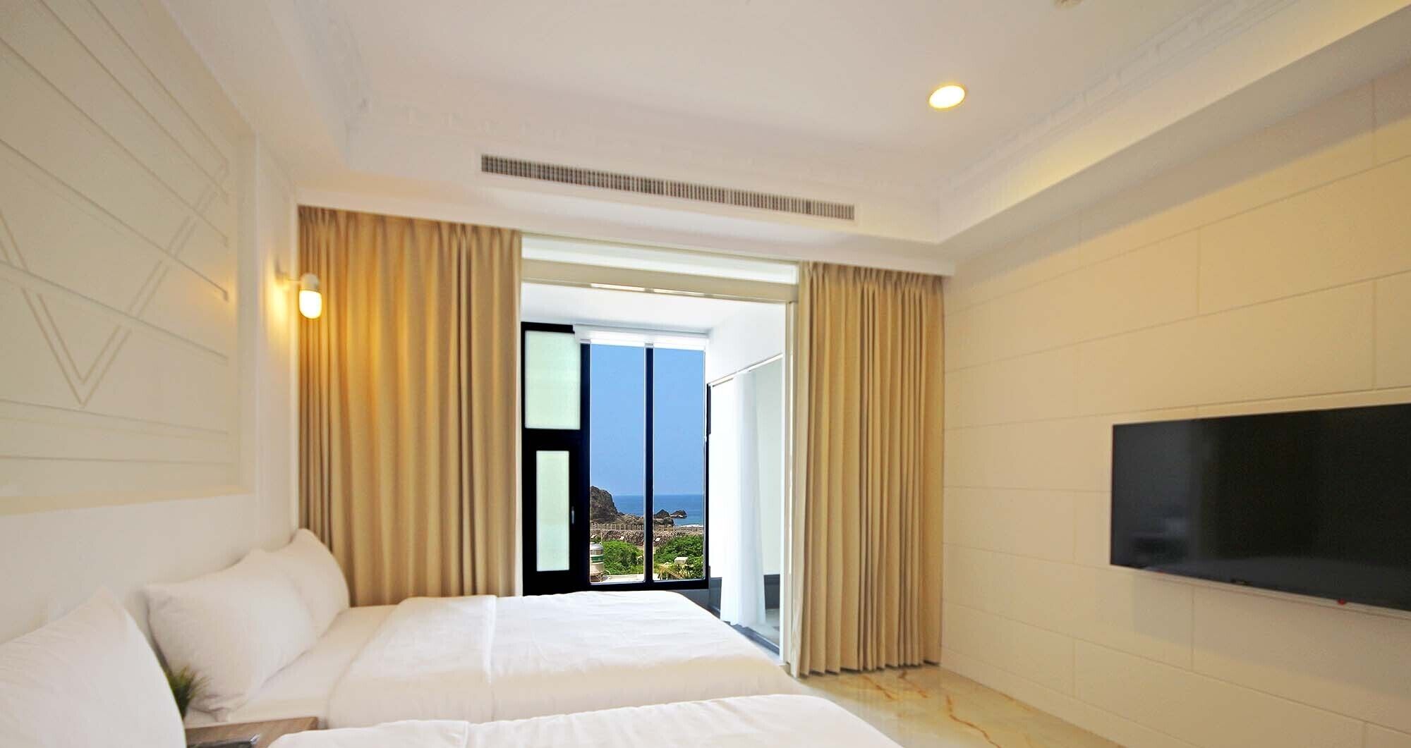 A hotel room with a bed, a TV, and a view of the sea through a sliding door.