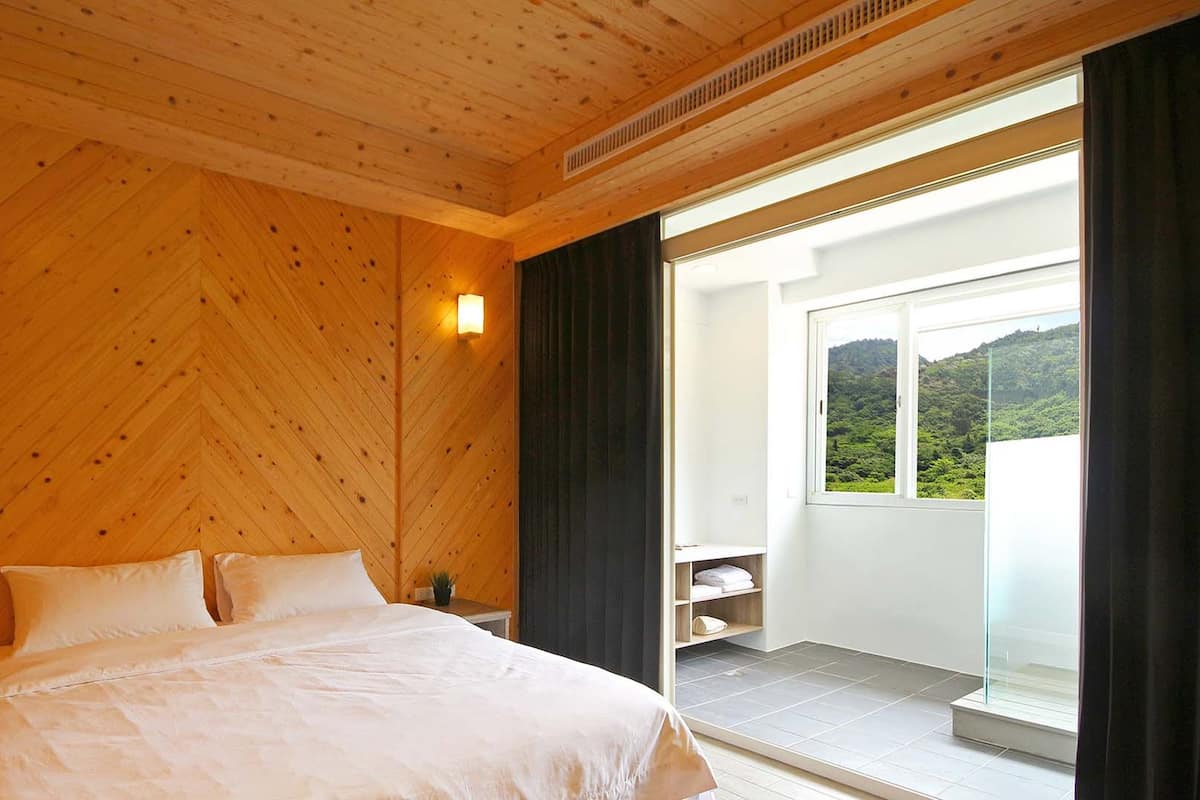 A bedroom with a wooden ceiling, a bed with white bedding, a bedside table, a nightstand, a small potted plant, a wall-mounted light, and a sliding glass door leading to a balcony with a view of greenery.