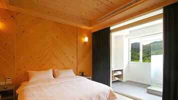 A bedroom with a wooden ceiling, a bed with white bedding, a bedside table, a nightstand, a small potted plant, a wall-mounted light, and a sliding glass door leading to a balcony with a view of greenery.
