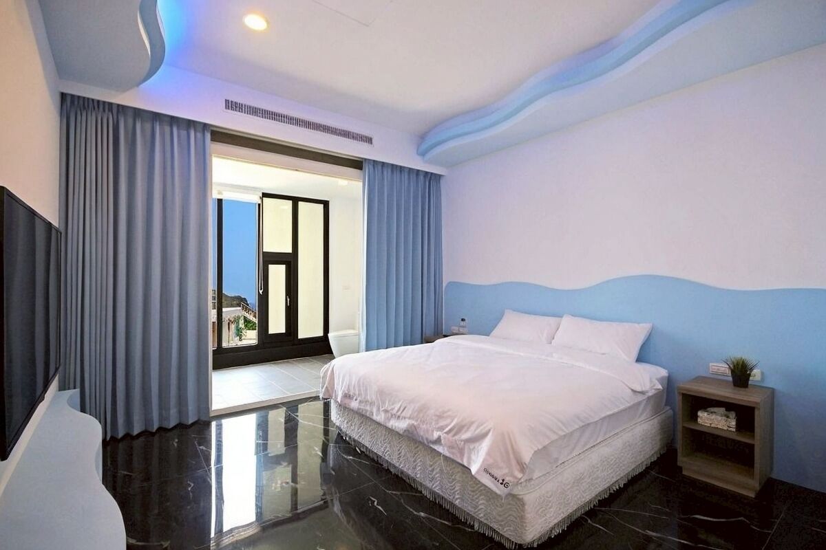 Une chambre d’hôtel moderne avec un grand lit, une table de chevet, une petite plante et une vue sur l’extérieur, visible à travers une porte vitrée.