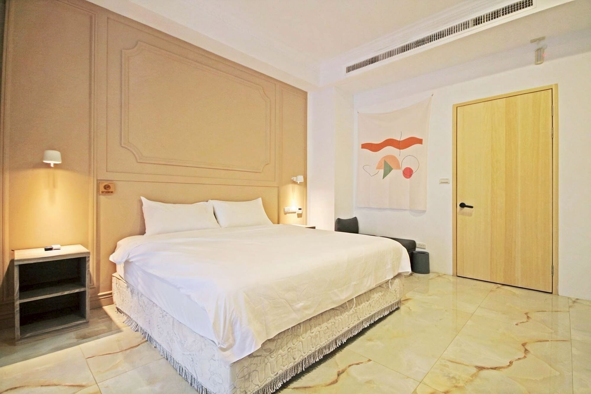 A hotel room with a large bed, a nightstand, a lamp, a door, and a wall with a decorative piece.