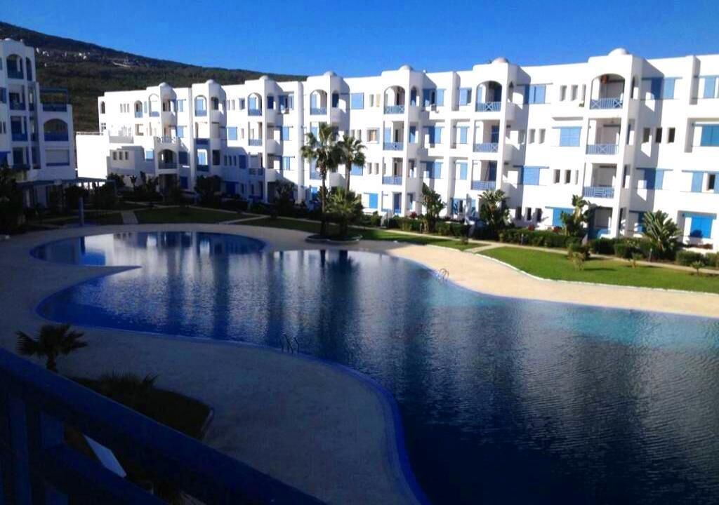 A resort with a large swimming pool, white buildings with blue accents, and a hilly landscape in the background.
