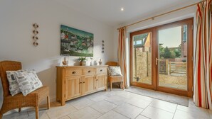 A spacious room with a large sliding glass door leading to an outdoor area, a wooden cabinet with drawers, a wicker chair, and a painting on the wall. - The Moorings, Wells-next-the-Sea, Norfolk (Wells-next-the-Sea)