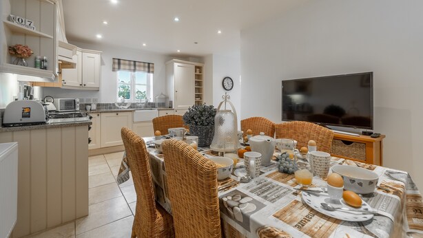 A modern kitchen with white cabinetry, a dining table set with breakfast items, wicker chairs, and a television on a stand. - The Moorings, Wells-next-the-Sea, Norfolk (Wells-next-the-Sea)