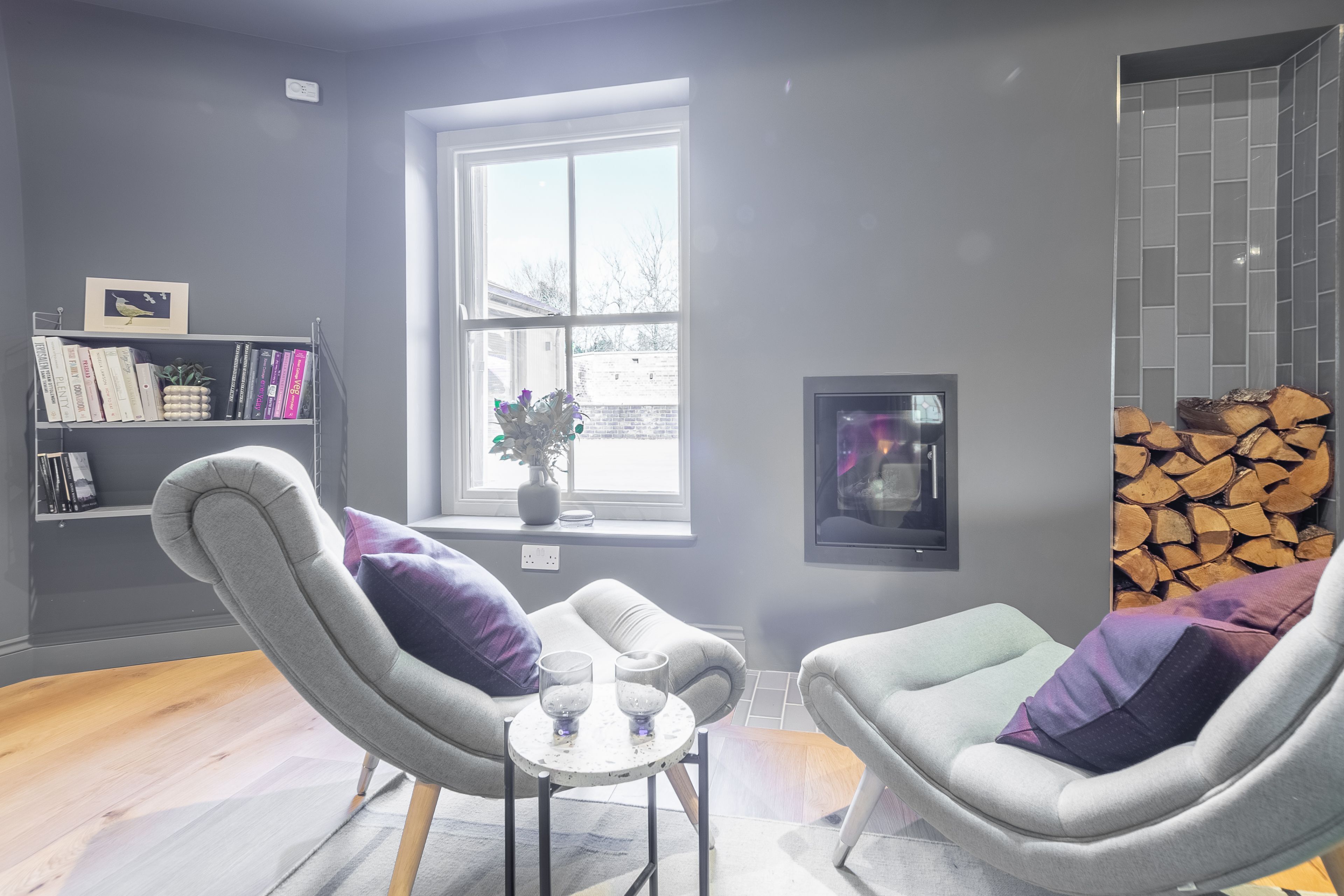 A modern living room with grey upholstered chairs, a small round table with glasses, a bookshelf, a window with a view, and a fireplace.