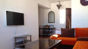 A living room with a flat-screen TV, a glass-top coffee table, a sofa with orange cushions, and a dining area with a mirror and chairs. - Residence Java Rasmile 2846 (Mdiq)