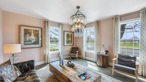 A spacious living room with a large window, a chandelier, a leather sofa, a wooden coffee table, and a framed painting.