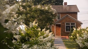 A wooden house with a brick chimney, surrounded by lush greenery and white flowering bushes.